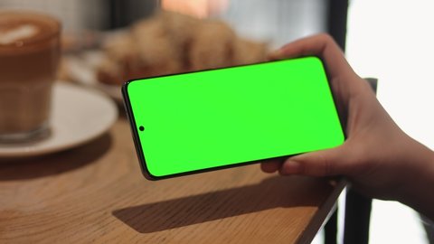 Back View of Female is scrolling, swiping her Smartphone with Green Screen, Chroma Key. Young Woman is Using Cell Phone at Restaurant, holding in Horizontal Position. Surfing Social Media.
