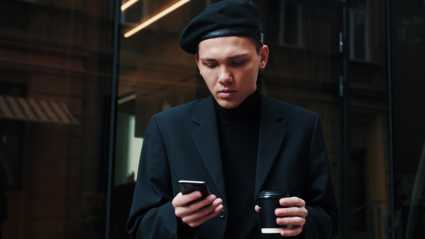 Smart casual dressed Office Worker is drinking Coffe at the Break. Attractive Man is standing near modern big Office and browsing his Smartphone. Office Workers. People. Business. Phones.