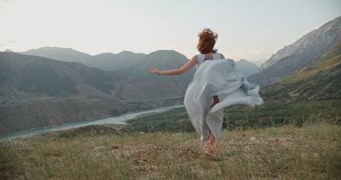 Young beautiful girl with red hair wearing white dress running on top of a mountain facing wind blowing her hair and dress and smiling - freedom, adventure, harmony 4k footage