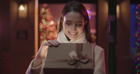 A young beautiful woman is opening a gift box, light shines on her face. She wears a white coat and she's happy. Christmas lights on the background.