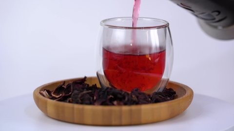 Red tea in a glass teapot. Hibiscus red tea pouring into a transparent glass close up view. Leaves of hibiscus near teacup in wooden stand. Free space for text. Footage for 
product advertising.