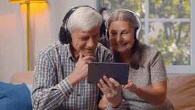 Portrait of happy elderly man and woman in headphones watching video on tablet pc at home. Smiling elderly couple in wireless headset surfing internet on digital tablet relaxing on couch