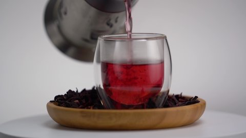 Red tea in a glass teapot on white background. Hibiscus red tea pouring into a transparent double wall glass.  Tea cup with herbal tea rotating on wooden board.