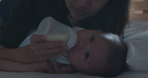 Cute asian baby girl drinking milk from baby bottle. Mother feeding daughter infant from bottle in bedroom before bedtime in slow motion shot.