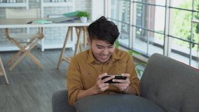 Happy emotion asian man is playing video game on sofa using modern smartphone.