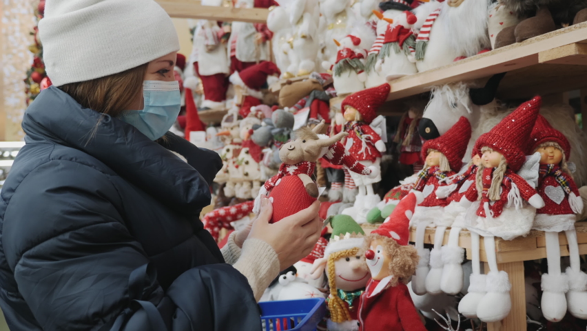A young woman in a medical mask shopping at the Christmas fair in the mall. Buying knitted toys for the holiday