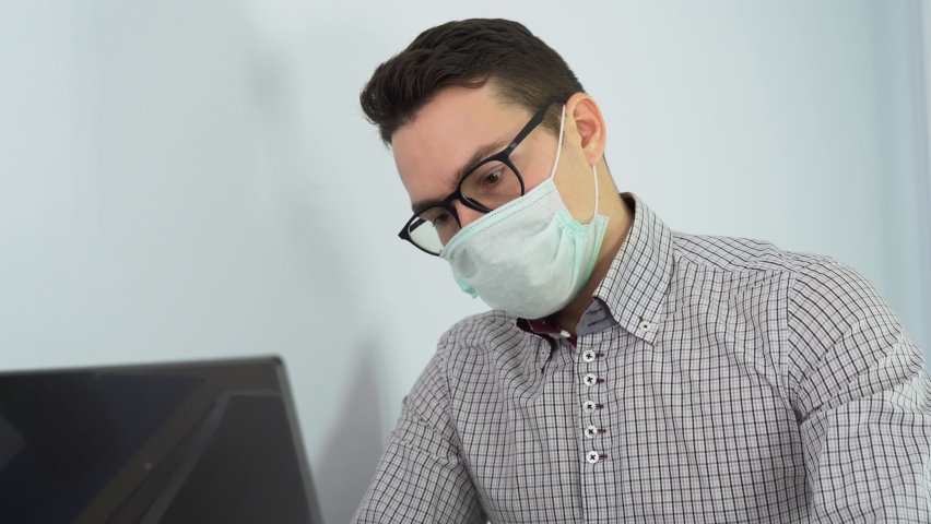 Alone man freelancer sitting in white room, workplace wear black glasses, medical face mask, latex gloves for not infect people around, use computer, typing on laptop keyboard, constantly coughing. 