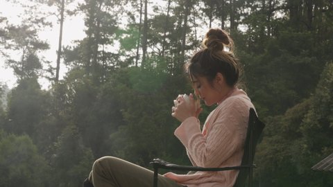 Young Asian woman sitting on outdoor balcony drinking a cup of hot coffee for breakfast in the morning. Beautiful female relax and enjoy outdoor activity lifestyle in nature on summer travel vacation.
