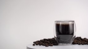  Black coffee in transparent, glass cup. Rotating video with glass and black coffee beans on white background. Empty place for text.  
Marketing, advertising use. Studio light.