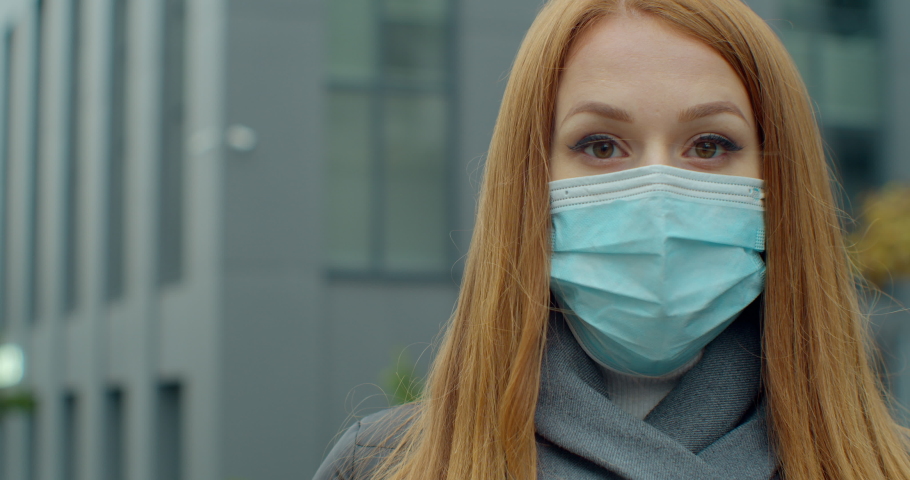 Close up portrait of a beautiful redhead young woman wearing protective medical face mask and standing on the street. Safe woman practicing social distancing, quarantine. | Shutterstock HD Video #1062544744