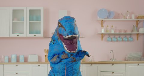 Funny dancing of blue dinosaur at pink kitchen. Comical movements of person in dino mascot. Slow motion fun dance.