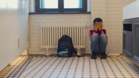 Depressed african child abandoned in lavatory and leaning against wall. Young afro boy sitting alone with sad feeling at school. Bullying, discrimination and racism concept