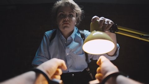 Female police officer with badge interrogating criminal and smoking cigarette. Close up of arrested man in handcuffs with woman detective turning lamp into his face