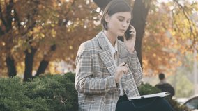Young beautiful female lawyer sitting on street talking on phone and making notes in her diary. Businesswoman working outdoor