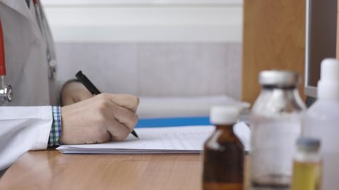 Male doctor doing paperwork in the office. Young therapist filling medical form or writtining a prescription for patient sitting at the desk. Health care and insurance concept.