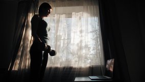 Silhouette of young girl pumping muscles with dumbbells watching tutorial on laptop. Woman doing sports and fitness training at home during covid 19 quarantine. Healthy lifestyle in pandemic time