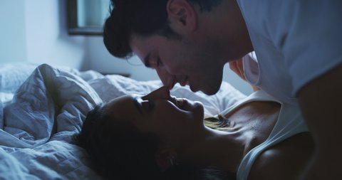 Cinematic shot of young just married couple in love wearing pajamas having fun and kissing on the bed in the bedroom in the early morning in a sunny day.