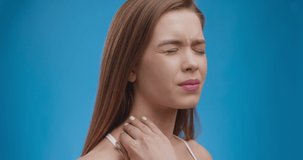 Young caucasian lady suffering from acute neck pain, massaging and stretching sore muscles, blue studio background, slow motion