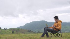 Asian man sitting on outdoor chair using laptop computer for online working video conference with drinking hot coffee on the mountain. Handsome guy enjoy outdoor lifestyle in autumn holiday vacation.