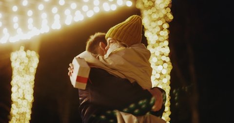 Young woman meeting up with boyfriend on birthday christmas evening party hugging tightly together swinging on the street. Illumination. Beautiful love couple.