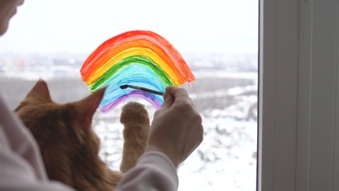 Young woman sitting with cat and painting rainbow on window during Covid-19 quarantine at home in winter. Stay at home Social media campaign for coronavirus prevention.