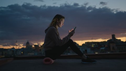 Side View of Young Pretty Woman Sitting on Yoga Mat on Roof Terrace at Sunset and Using Smartphone for Communication, Entertainment or Study. Modern Technology Concept. Slow Motion Cinematic Shot