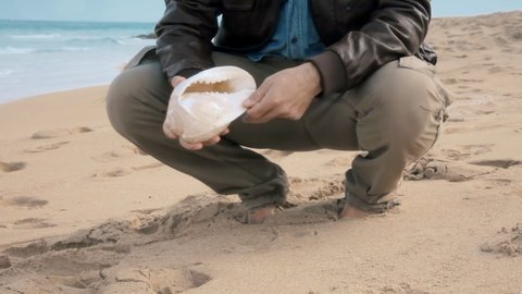 A man slowly walking on a sandy beach, picking up a seashell, cleaning it, listening to the sounds by putting it near his ear, leaving it where he found it. Slow zoom in.
