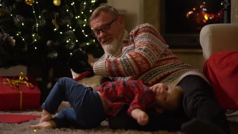 Elderly man and kid celebrate christmas, happy family. Grandfather and grandson and the dog have fun lying on the carpet near the Christmas tree. Family traditions