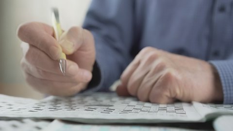 older man with yellow pen in wrinkled hand writing letters in croosword or sudoku during coronavirus quarantine