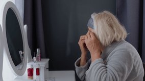 Old senior woman grandmother taking care of skin near eyes and wrinkles, putting makeup on, looking into a mirror. Elderly grandma doing make-up procedures at home. Cosmetics for retired pensioners