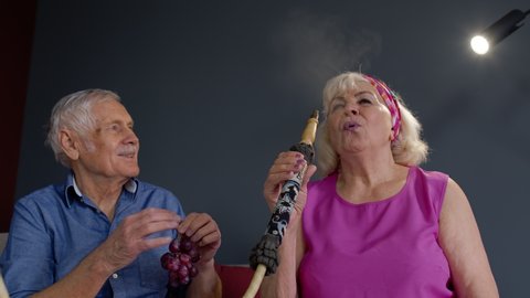 Happy stylish senior couple smoking hookah at home. Modern trendy elderly grandparents celebrating their anniversary relaxing having fun together enjoy care love tenderness. Slow motion. 6k downscale