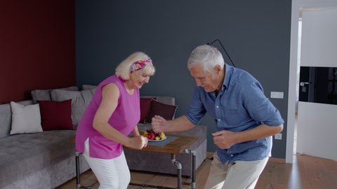 Happy old senior couple dancing celebrating retirement anniversary in modern living room at home. Grandmother and grandfather having fun dance enjoying relationship milestone celebration. 6k downscale