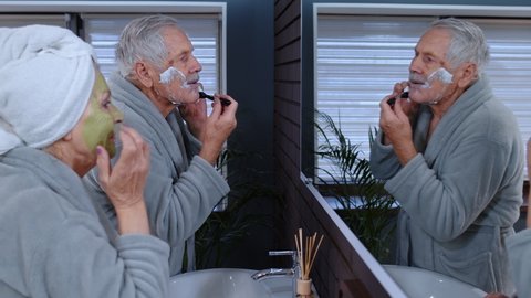 Old senior couple man and woman doing morning hygiene and looking into a mirror. Attractive elderly grandmother applying facial green mask and grandfather shaving with manual razor blade at bathroom