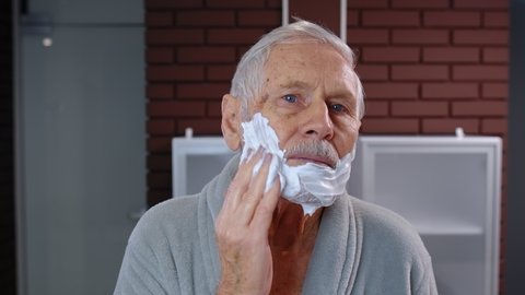 Attractive old senior man grandfather in bathrobe applying foam to face for shaving and looking into mirror. Handsome elderly grandpa doing morning skin hygiene after shower at bathroom home. POV shot
