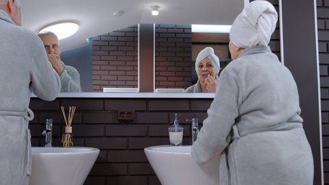 Cheerful old senior couple grandparents man and woman brushing teeth and looking into a mirror. Elderly grandmother and grandfather doing morning hygiene at luxury bathroom at home. Slow motion