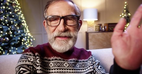 Close up of cheerful smiling old male pensioner in glasses chatting on video call in room with glowing decorated xmas tree. POV of grey-haired man speaking on video online congratulating with holidays