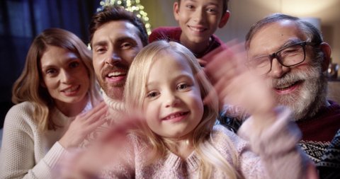 POV f joyful lovely Caucasian family with kids gathered together in cozy room with glowing xmas tree and video chatting online sending holiday greetings to friends Happy holidays concept