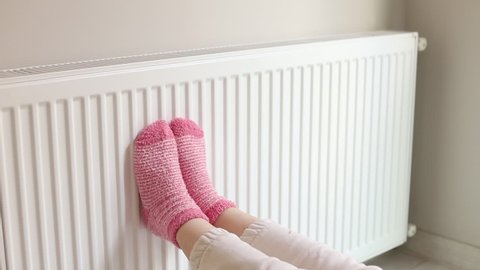 Woman is warm knitted woolen socks near a home heater in cold winter time. Using heater at home in winter.
