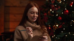 Close-up of happy dreamy young woman writing wish list for new year on background of Christmas tree at cozy dark living room with festive interior. Cheerful lady writing xmas letter to Santa Claus. - Powered by Shutterstock - Get 15% off with code: PIKWIZARD15