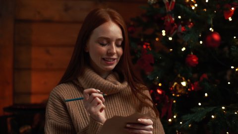 Close-up of happy dreamy young woman writing wish list for new year on background of Christmas tree at cozy dark living room with festive interior. Cheerful lady writing xmas letter to Santa Claus.