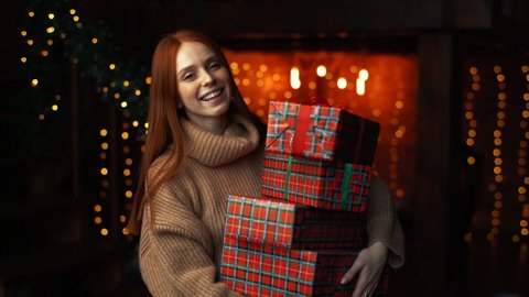 Happy young woman holding many beautiful Christmas gift boxes on background of xmas lights at cozy dark room. Cheerful lady drops box with gift, tries to catch and looks at camera enthusiastically.
