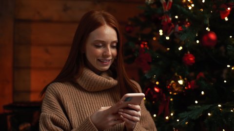 Portrait of happy young woman wearing winter sweater typing online message using mobile phone on background Christmas tree with bright lighting at cozy dark living room with festive interior.