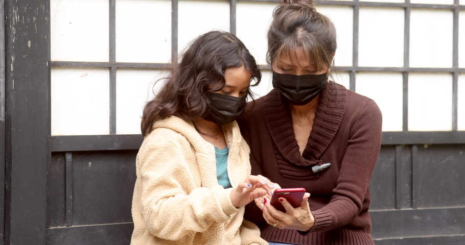Attractive woman sitting with young girl, both wearing covid masks for coronavirus pandemic, playing game on cell phone seated on bench. Outdoor background of japanese black and white squares.