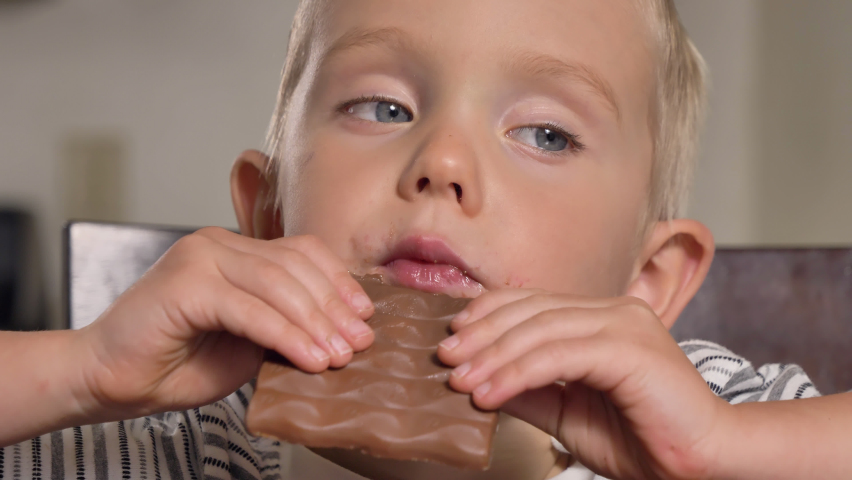 Child (little Caucasian boy) eating chocolate, biting candies, looking at camera. Unhealthy food, childhood concept. Indoor 