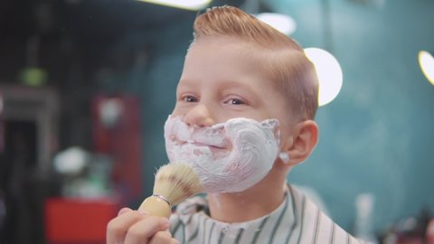Little boy smears shaving foam on his face with a shaving brush. Hike to the barbershop