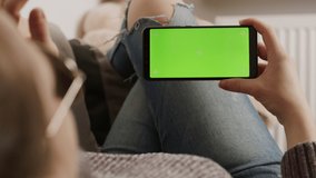 Young woman holding smartphone with green screen while resting on the sofa and waving. Video call. High quality 4k footage
