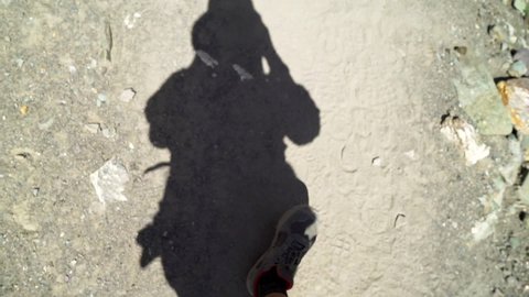 POV shot filming own shadow as walking on a dirt road, a hiker in nature, with the feet in the frame, looking down. 