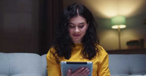 Beautiful Caucasian female sitting on sofa and using digital tablet in living room. Good-looking young woman holding gadget and looking at camera at home in evening. Technology concept.