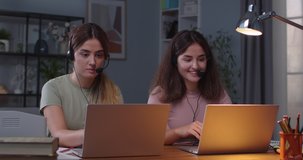 Joyful Caucasian twin sisters sitting in head sets at table in room and speaking on video conference on laptops Females having online lesson while working on computers at home Distance work E-learning