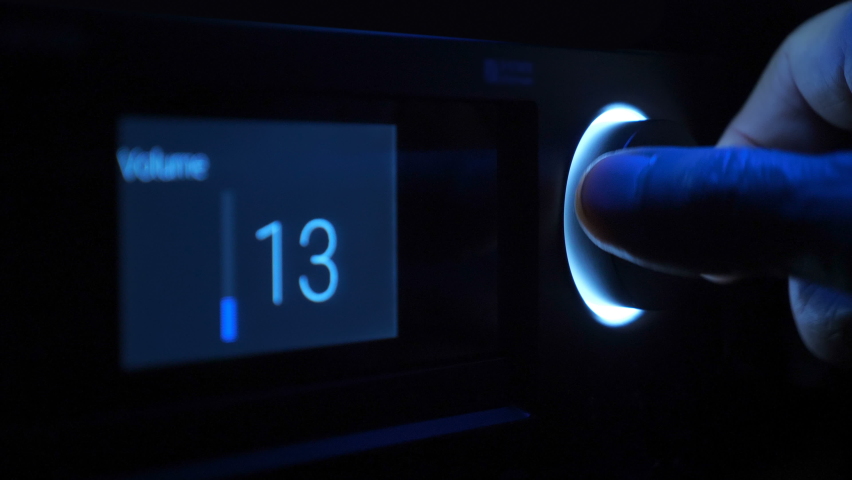 In the dark, the hand turns the volume control, illuminated by blue light on the front panel of the black stereo equipment. The display shows the music volume level. Closeup | Shutterstock HD Video #1062596515
